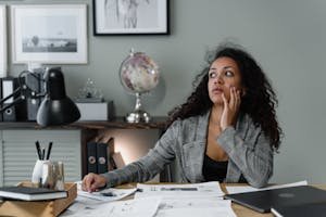 A businesswoman in a plaid blazer at her desk, thinking amid documents and gadgets in an office setting.